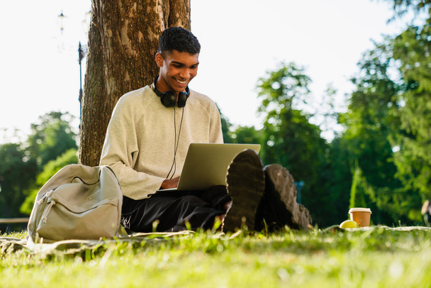 Junger fröhlicher afrikanischer Mann in Freizeitkleidung arbeitet am Laptop, während er im grünen Park sitzt - Foto, Bild