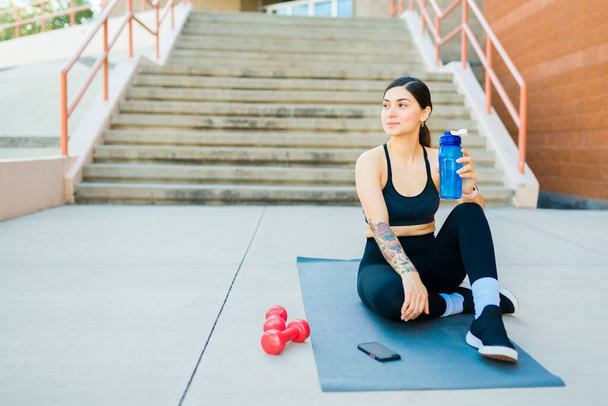 Souriant jeune femme en forme avec une bouteille d'eau regardant heureux de travailler à l'extérieur sur un tapis d'exercice - Photo, image