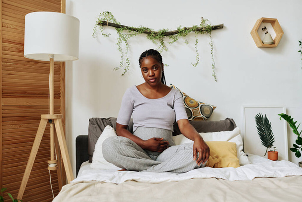 Yatak odasında boş zamanlarında yatakta oturmuş kameraya bakan Afrikalı hamile bir kadının portresi. - Fotoğraf, Görsel