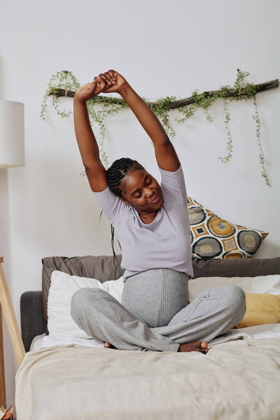African pregnant woman sitting on bed and stretching while waking up in the morning - Photo, Image