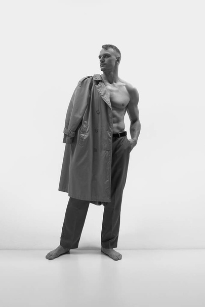 Atractivo hombre musculoso de pie y sosteniendo el abrigo en el hombro sobre fondo blanco del estudio. Blanco y negro. Concepto de moda masculina, estilo, estética corporal, belleza, salud masculina, emociones - Foto, Imagen