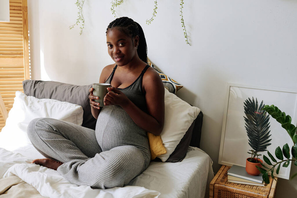 Yatakta çay içerken kameraya gülümseyen Afrikalı hamile kadın portresi. - Fotoğraf, Görsel