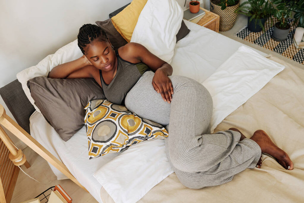 High angle view of African young girl looking at her pregnant belly while relaxing on cushions in bed - Фото, изображение