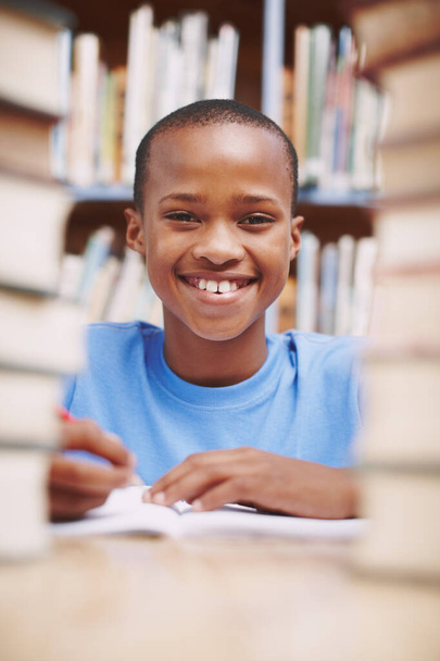 Tuo leveä hymy kertoo, että hän haluaa oppia. Afrikkalainen amerikkalainen poika hymyilee sinulle kirjastossa kirjapinon välissä. - Valokuva, kuva
