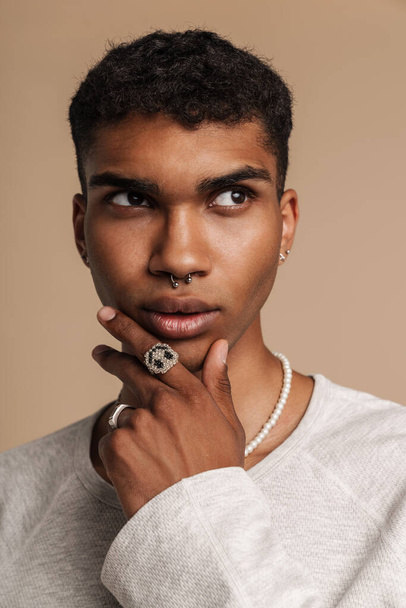 Young black man wearing rings looking aside while holding his chin isolated over beige background - Foto, Bild