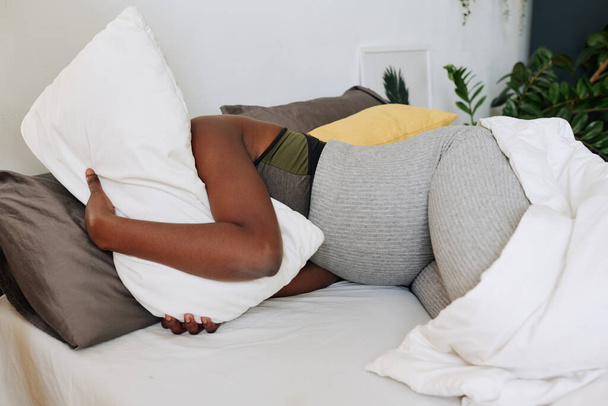 Afrykańska kobieta w ciąży obejmująca poduszkę i cierpiąca z powodu bólu leżącego na łóżku podczas porodu - Zdjęcie, obraz