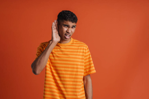 Portrait of young handsome confused african man with hand near ear trying to hear something looking at camera, whila standing over isolated orange backgr - Fotó, kép