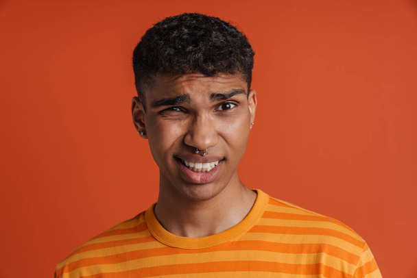 Portrait of young handsome confused african man with piercing looking at camera , while standing over isolated orange background - Foto, imagen