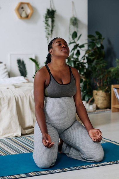 Afrikkalainen raskaana oleva nainen istuu harjoitusmatolla ja hengittää syvään meditaation aikana kotona - Valokuva, kuva