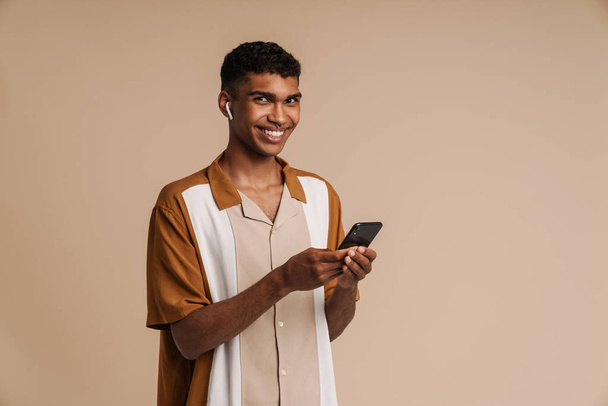 Young african handsome smiling man in wireless headphones holding phone and looking at camera , while standing over isolated beige background - Фото, изображение