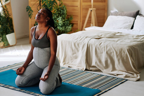 African pregnant girl sitting on exercise mat and meditating with her eyes closed in bedroom - Photo, Image
