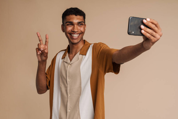 Mladý africký hezký usmívající se muž brát selfie s telefonem a ukazuje vítězství gesto, zatímco stojí nad izolované béžové pozadí - Fotografie, Obrázek