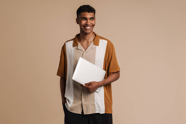 Young handsome smiling african man holding laptop looking aside , while standing over isolated beige background - Φωτογραφία, εικόνα