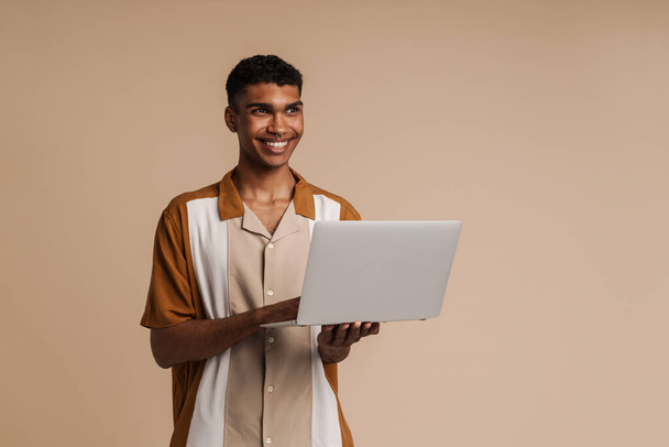 Young handsome smiling african man with piercing holding laptop and looking aside , while standing over isolated beige background - Fotoğraf, Görsel