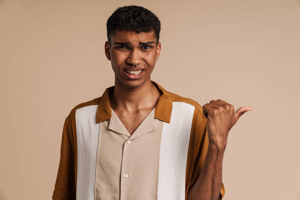 Young handsome confused african man with piercing pointing aside with thumb finger and looking at camera , while standing over isolated beige background - Foto, afbeelding