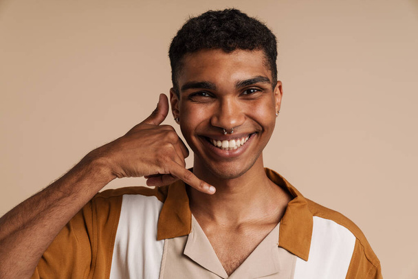 Young handsome smiling happy african man with piercing doing phone gesture with hand and fingers and looking at camera, while standing over isolated beige background - Fotó, kép