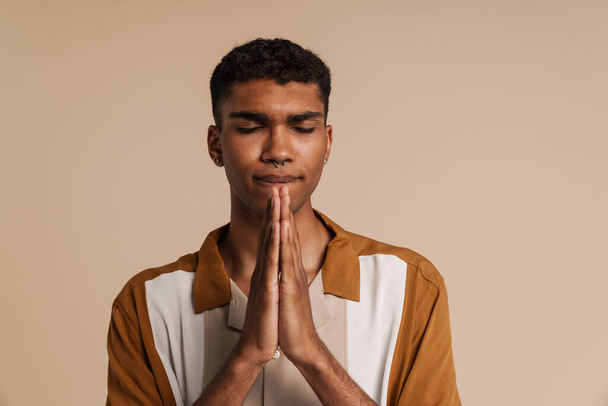Young handsome african man with closed eyes doing pray gesture with folded hands, while standing over isolated beige background - Foto, Imagem