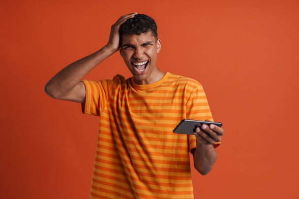 Young handsome disappointed african man with opened mouth touching head, holding phone and looking at camera , while standing over isolated orange background - Photo, image