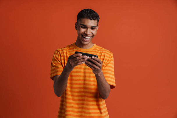 Young handsome smiling happy african man playing mobile game on phone, while standing over isolated orange background - Photo, Image