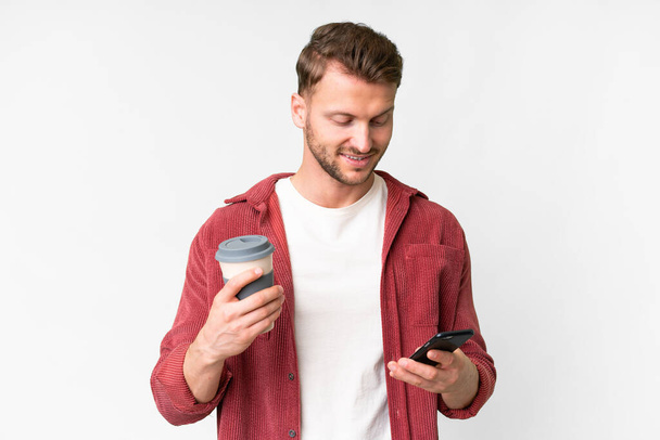 Fiatal jóképű kaukázusi férfi izolált fehér háttér kezében kávé elvitelre és egy mobil - Fotó, kép