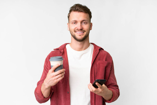Mladý pohledný kavkazský muž přes izolované bílé pozadí drží kávu odnést a mobilní - Fotografie, Obrázek
