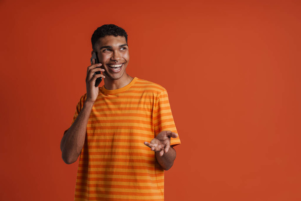 Giovane bell'uomo africano felice sorridente con piercing parlare al telefono e guardare da parte, mentre in piedi sopra isolato sfondo arancione - Foto, immagini