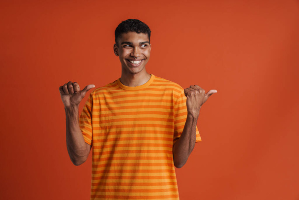 Young handsome smiling happy african man with piercing, looking and pointing aside with both thumbs , while standing over isolated orange background - Фото, зображення