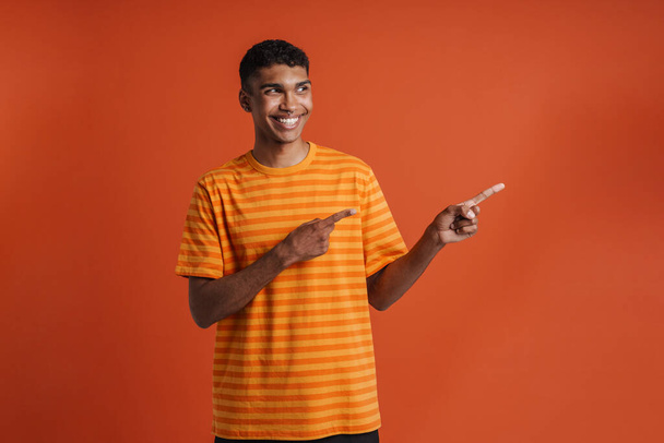Young handsome smiling happy african man with piercing looking and pointing aside with both hands , while standing over isolated orange background - Foto, Imagen