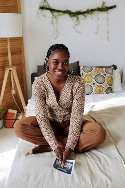 Portrait of African young woman sitting on bed and smiling at camera, she learning about her pregnancy - Photo, Image
