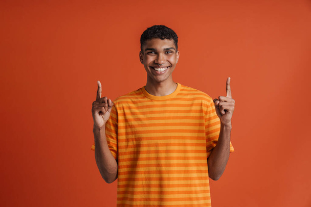 Young handsome smiling happy african man with piercing pointing upward with both hands and looking at camera, while standing over isolated orange background - Foto, Imagen