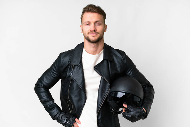 Joven hombre caucásico con un casco de moto sobre fondo blanco aislado posando con los brazos en la cadera y sonriendo - Foto, Imagen