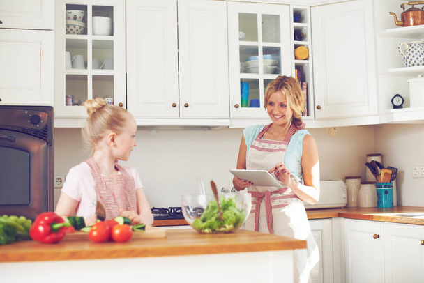 Вы делаете большую работу, следуя этому рецепту. Маленькая девочка делает салат на кухне со своей матерью, стоящей рядом, используя цифровой планшет - Фото, изображение