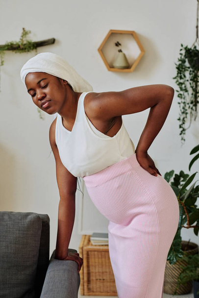 Africana musulmana embarazada sintiendo dolor en la espalda durante el parto, ella se apoya en el sofá con los ojos cerrados - Foto, imagen