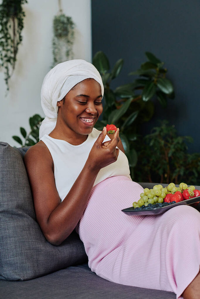 Mujer musulmana africana en hijab sentada en un sofá y comiendo frutas frescas durante su embarazo - Foto, imagen