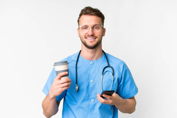 Jeune infirmier homme sur fond blanc isolé tenant du café à emporter et un mobile - Photo, image