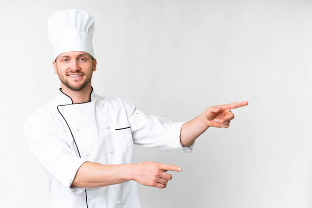 Молодой кавказский повар на изолированном белом фоне, указывая пальцем в сторону и представляя продукт - Фото, изображение