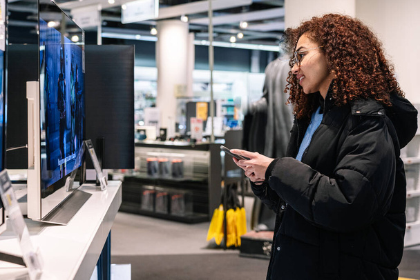 Side view of young African American female in casual clothes smiling and browsing smartphone while standing near TV in electronic store - Фото, зображення