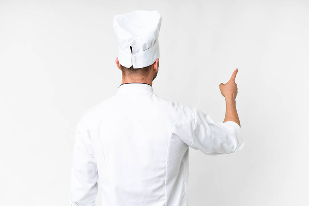 Mladý kavkazský kuchař přes izolované bílé pozadí ukazuje zpět s ukazováčkem - Fotografie, Obrázek