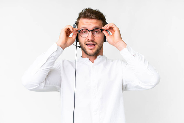 Telemarketer hombre caucásico trabajando con un auricular sobre fondo blanco aislado con gafas y sorprendido - Foto, imagen
