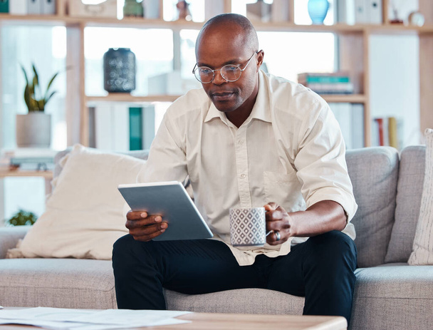 Tablet, coffee and relax with a black man on the sofa, sitting in the living room of his home or office. Business, tech and research with a male employee reading an online article while on a break. - Foto, Imagem