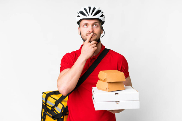 Young delivery man taking a takeaway food over isolated white background having doubts while looking up - Photo, Image