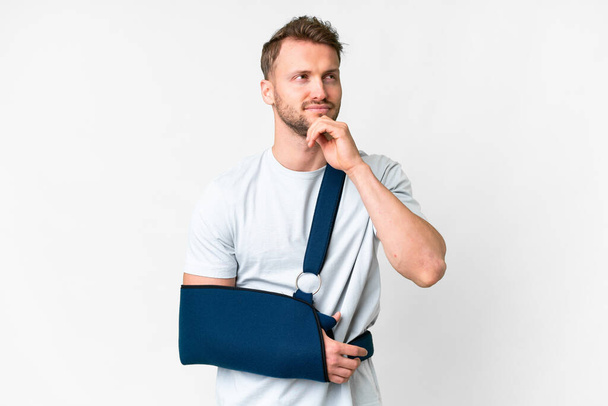 Young caucasian man with broken arm and wearing a sling over isolated white background and looking up - Foto, Imagem