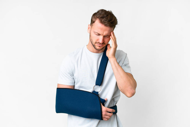 Young caucasian man with broken arm and wearing a sling over isolated white background with headache - Photo, Image