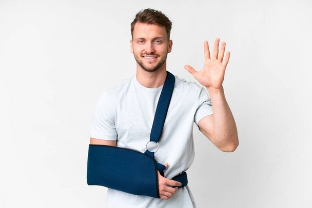 Young caucasian man with broken arm and wearing a sling over isolated white background saluting with hand with happy expression - Φωτογραφία, εικόνα