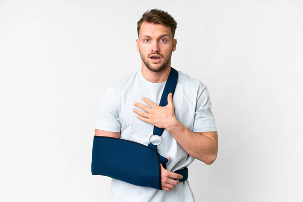 Mladý kavkazský muž se zlomenou rukou a na sobě prak přes izolované bílé pozadí překvapen a šokován při pohledu vpravo - Fotografie, Obrázek