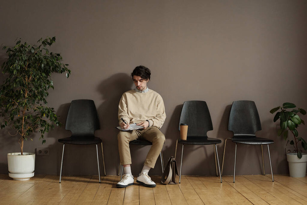 Young man sitting on chair in waiting room and filling form before job interview - Фото, изображение