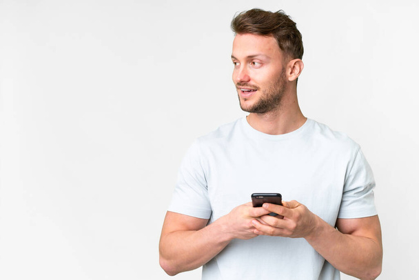 Молодой красивый белый мужчина на изолированном белом фоне с помощью мобильного телефона и глядя вверх - Фото, изображение