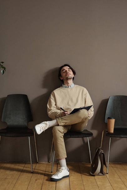 Młody człowiek z formularzem aplikacyjnym spoczywający na krześle z zamkniętymi oczami w oczekiwaniu na rozmowę kwalifikacyjną - Zdjęcie, obraz