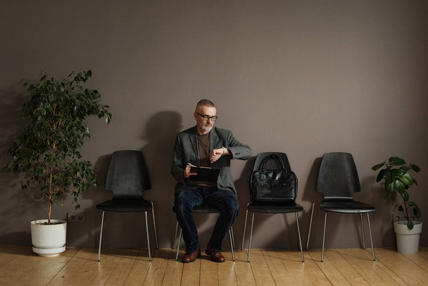 Reifer Mann mit Brille kontrolliert die Uhrzeit auf seiner Uhr, während er im Wartezimmer sitzt und das Formular ausfüllt - Foto, Bild