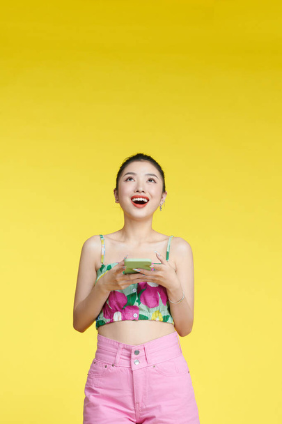 Giovane sorpreso donna divertente stupefatto indossando serbatoio superiore tenere utilizzando cellulare isolato su sfondo giallo  - Foto, immagini